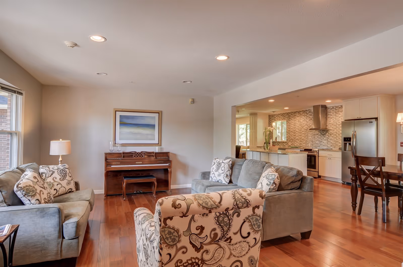Open-concept living room with sofas and a patterned armchair, a piano against the wall, and a view into the adjoining kitchen.