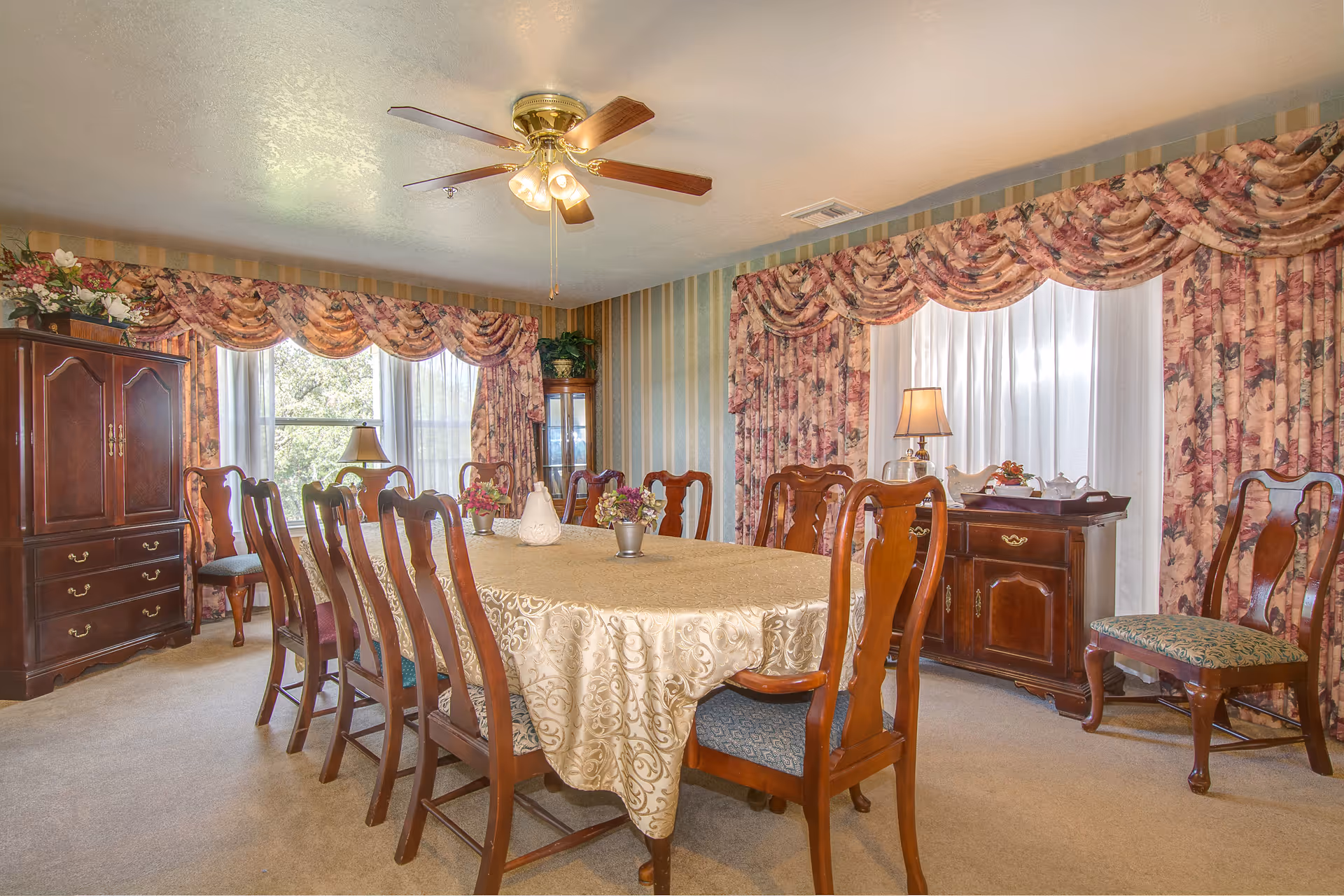 A traditional dining room with a long rectangular table covered with a beige patterned tablecloth surrounded by wooden chairs with upholstered seats. The room features floral curtains with valances on two large windows, a ceiling fan with lights, a wooden cabinet with drawers, a sideboard with a lamp and tea set, and decorative plants and flowers.