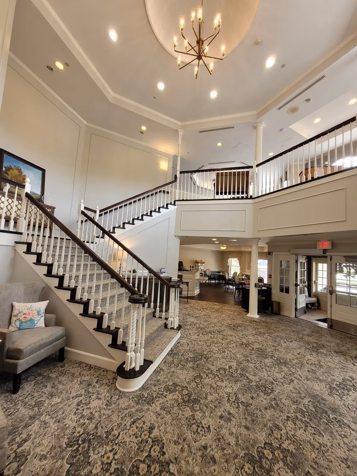 Interior view of a senior living facility lobby with a carpeted floor, a staircase with white railings and dark wood handrails leading to an upper level, a chandelier hanging from a high ceiling, a gray armchair with a floral pillow, and a seating area with tables and chairs in the background near large windows.