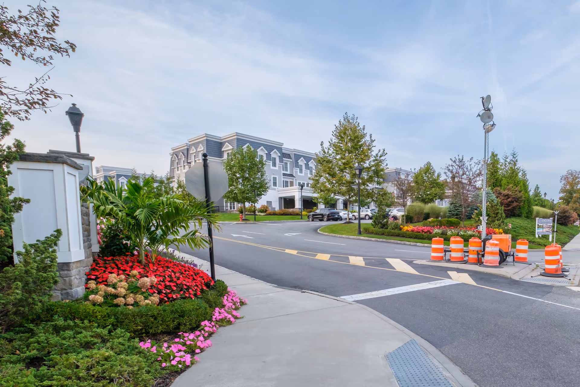 View of the entrance road to a senior living facility with landscaped flower beds, trees, and a large multi-story building in the background under a partly cloudy sky. Orange construction barrels and a tall light stand are visible near the road.