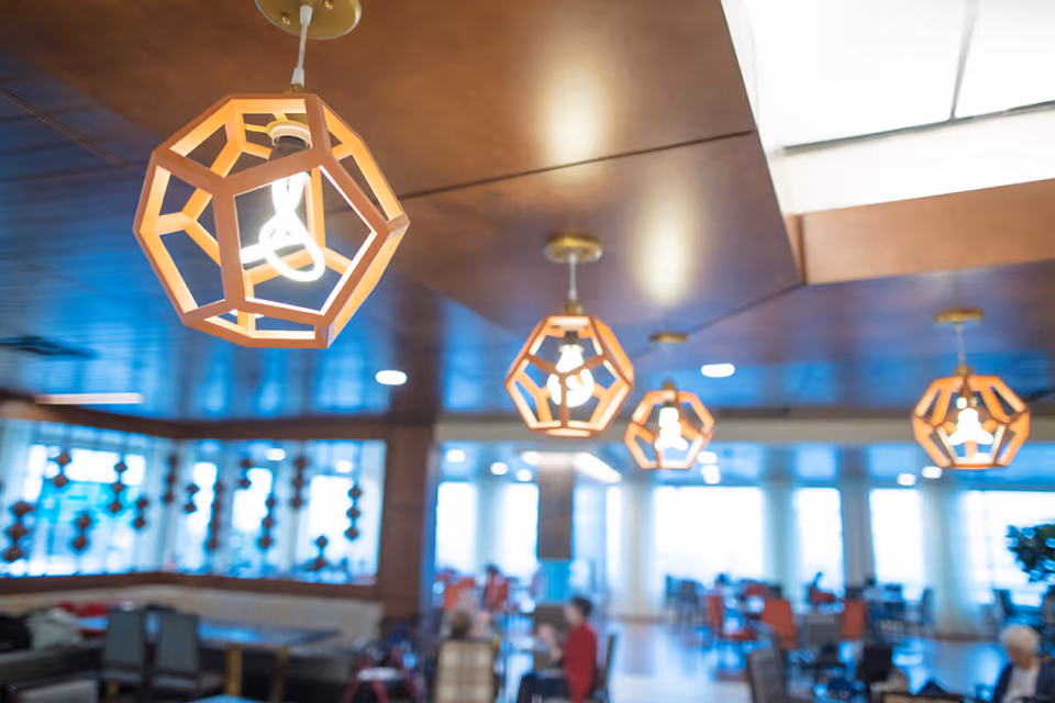 Geometric pendant lights hang from a wooden ceiling above a blurred communal dining area with tables and people.
