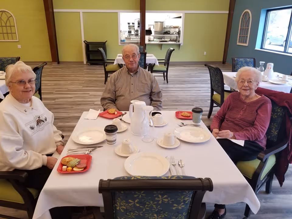 Three elderly people sitting around a dining table in a well-lit room with green and blue walls. The table is set with white plates, cups, and a white pitcher. Each person has a red tray with snacks and a cup of coffee. The room has wooden flooring and several tables and chairs in the background.
