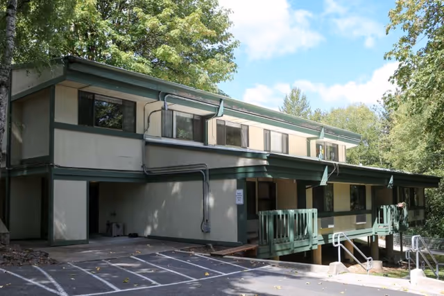Exterior view of a two-story building with beige walls and green trim surrounded by trees under a partly cloudy sky. The building has multiple windows and a small porch with green railings. There is a parking area with marked spaces in front of the building.