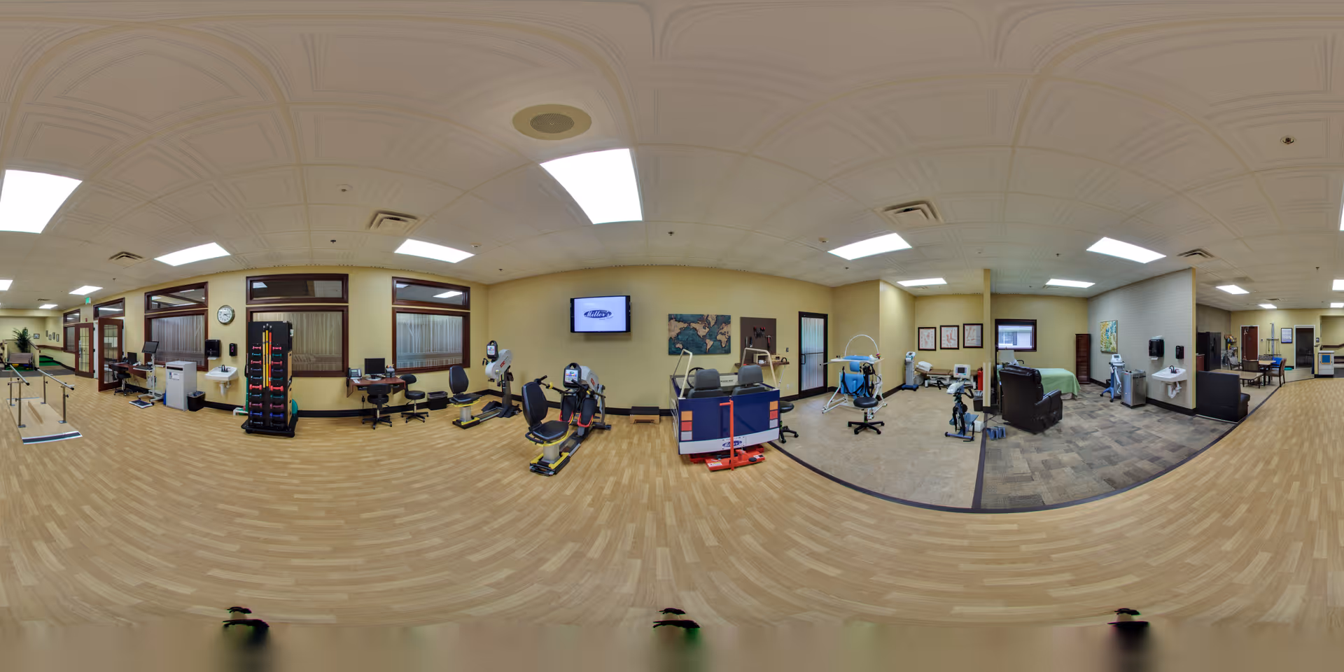 A spacious rehabilitation or physical therapy room with exercise equipment including stationary bikes, parallel bars, and therapy chairs. The room has wood-patterned flooring, beige walls, and ceiling lights. There are sinks, chairs, and therapy tools visible, along with a TV mounted on the wall and some artwork. The area appears clean and well-organized, designed for senior care physical therapy.