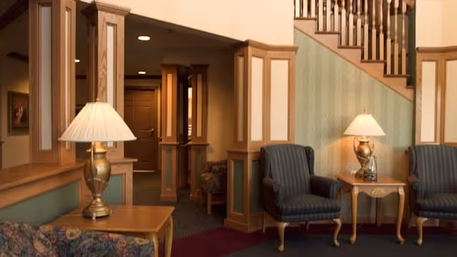 A cozy interior common area with wooden paneling and trim, featuring two blue upholstered armchairs with wooden legs, two wooden side tables each with a brass table lamp, a floral patterned sofa, and a staircase with wooden banisters in the background.