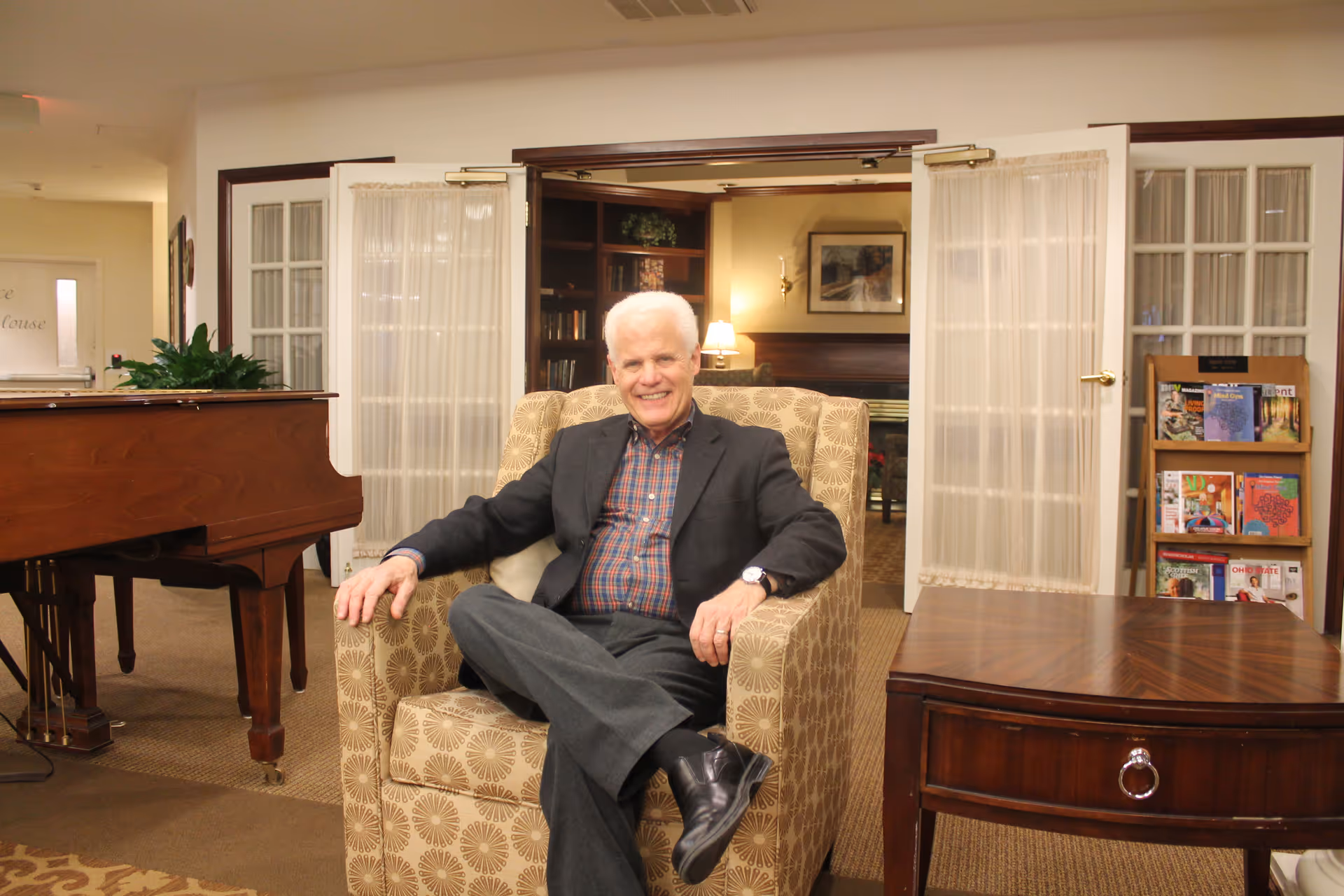 An elderly man with white hair, wearing a dark blazer and plaid shirt, sits comfortably in a patterned armchair in a warmly lit room. Behind him are glass-paneled double doors with sheer curtains, a wooden magazine rack filled with magazines, and a grand piano to the left. The setting appears to be a cozy common area or lounge in a senior living facility.