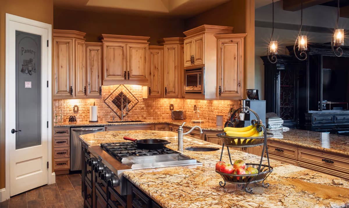 A spacious kitchen with wooden cabinets, granite countertops, a gas stove with a frying pan, a double-tiered fruit basket holding bananas and apples, a sink, and a pantry door with frosted glass labeled 'Pantry'. Warm lighting illuminates the backsplash and countertops.