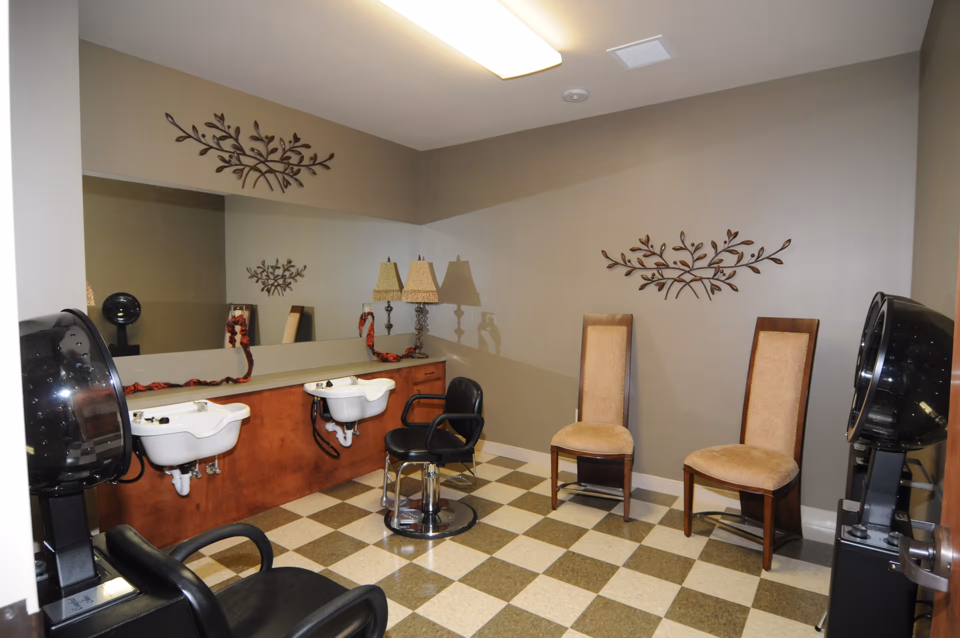 Interior of a hair salon area in a senior living facility with two white wash basins, black salon chairs, two beige upholstered chairs, a large mirror on the wall, decorative metal wall art, and a checkered floor.