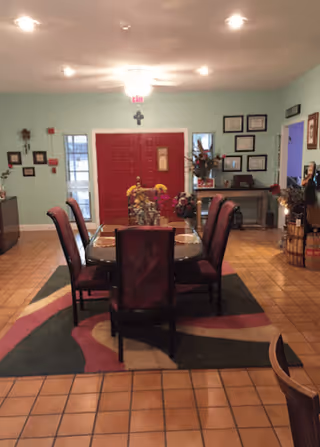 Interior view of a dining area with a rectangular table surrounded by six chairs. The floor is tiled with a patterned rug under the table. The walls are decorated with framed pictures and a cross above a red double door. There is a side table with flower arrangements and additional framed certificates or pictures on the wall.