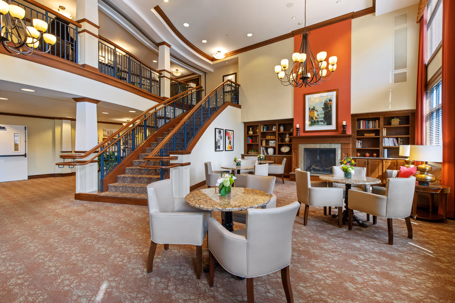 Spacious communal lounge with round tables and upholstered chairs, a grand staircase, fireplace, bookshelves, and chandeliers.