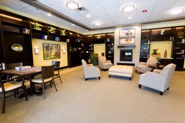 A spacious living room area with four striped armchairs arranged around a large ottoman in front of a fireplace. The walls feature built-in dark wood shelves with decorative items and plants. There is a table with chairs on the left side of the room, and the ceiling has recessed lighting and circular light fixtures.