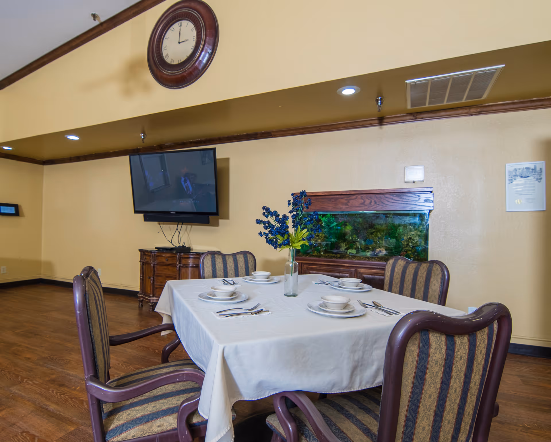 A dining area with a square table covered with a white tablecloth set for four people with plates, bowls, and cutlery. Four upholstered chairs surround the table. In the background, there is a wall-mounted flat-screen TV above a wooden cabinet and a large fish tank with plants inside. A large round clock is mounted high on the wall above the TV.