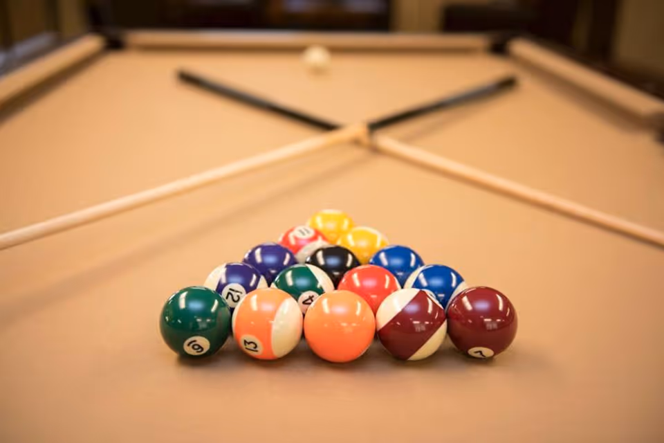 A close-up view of a beige pool table with a neatly racked set of colorful billiard balls in a triangle formation and two pool cues crossed in the background.
