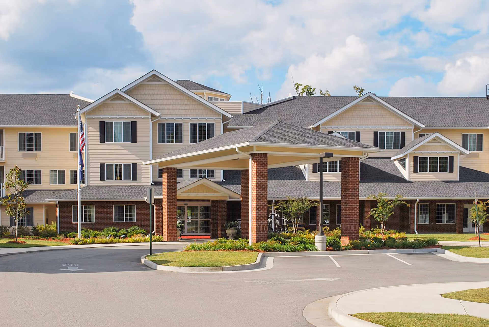 Front exterior view of Dogwood Terrace by Barclay House, a multi-story senior living facility with beige siding, dark shutters, and a covered entrance supported by brick columns. The building is surrounded by landscaped greenery and a paved driveway under a partly cloudy sky.