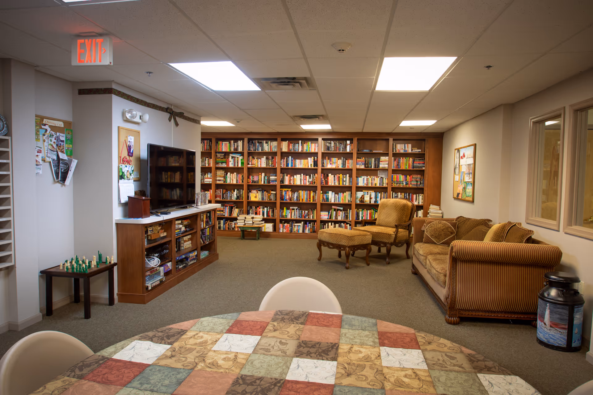 A cozy common room with floor-to-ceiling bookshelves, a TV and media console, upholstered chairs and a sofa, and a table with a patchwork tablecloth in the foreground.