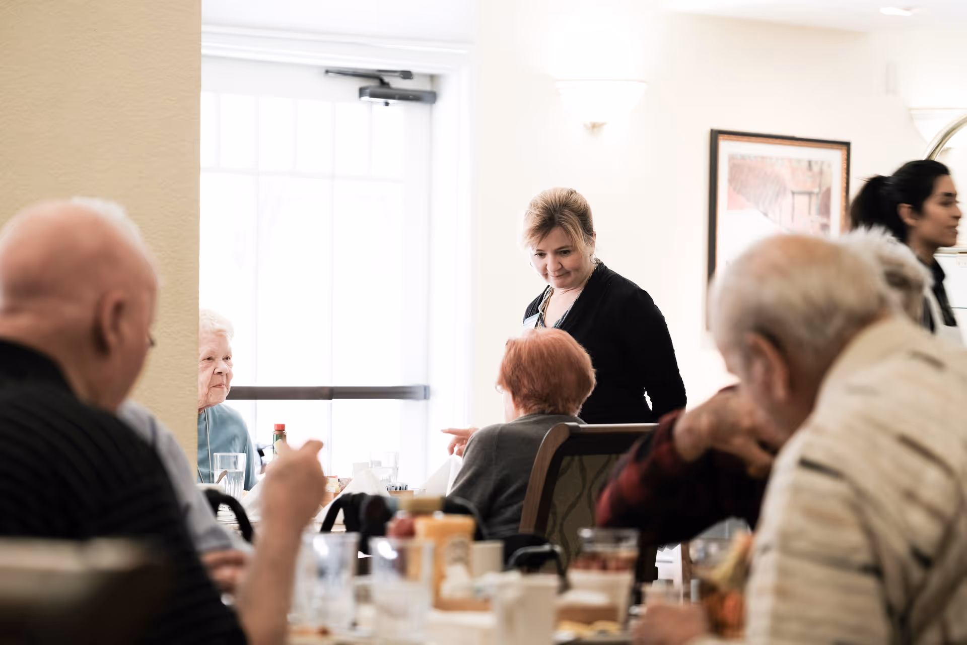 A group of elderly people sitting around a dining table in a well-lit room, with a caregiver standing and interacting with them. The table has various items including glasses, condiments, and plates. The background shows a door with a window and framed artwork on the wall.