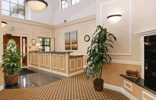 Reception area of Grossmont Gardens Memory Care with a front desk, potted plants, wall clock, framed artwork, and a door with exit sign above it. The area has beige walls with white trim and patterned carpet flooring.