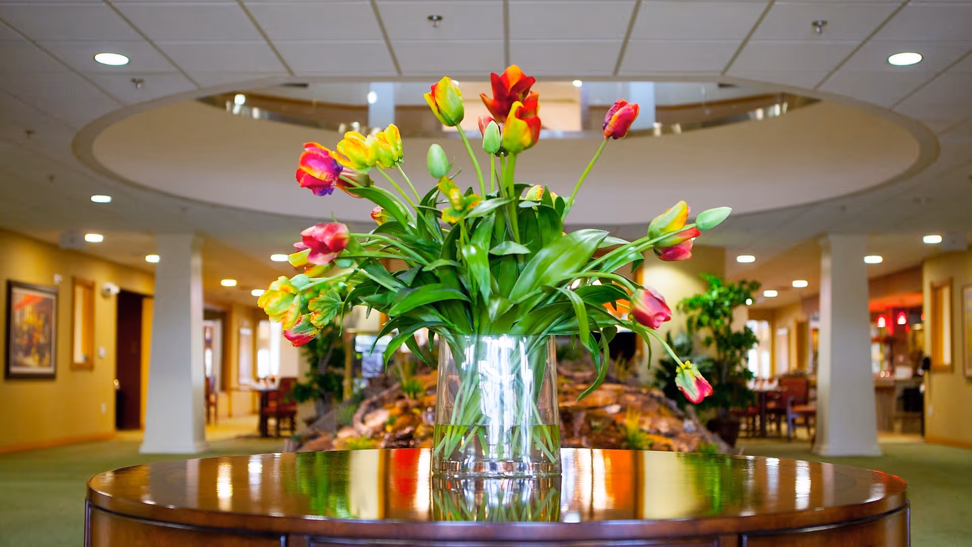A clear glass vase filled with colorful tulips, placed on a polished wooden round table in the center of a spacious, well-lit interior common area with plants and seating in the background.