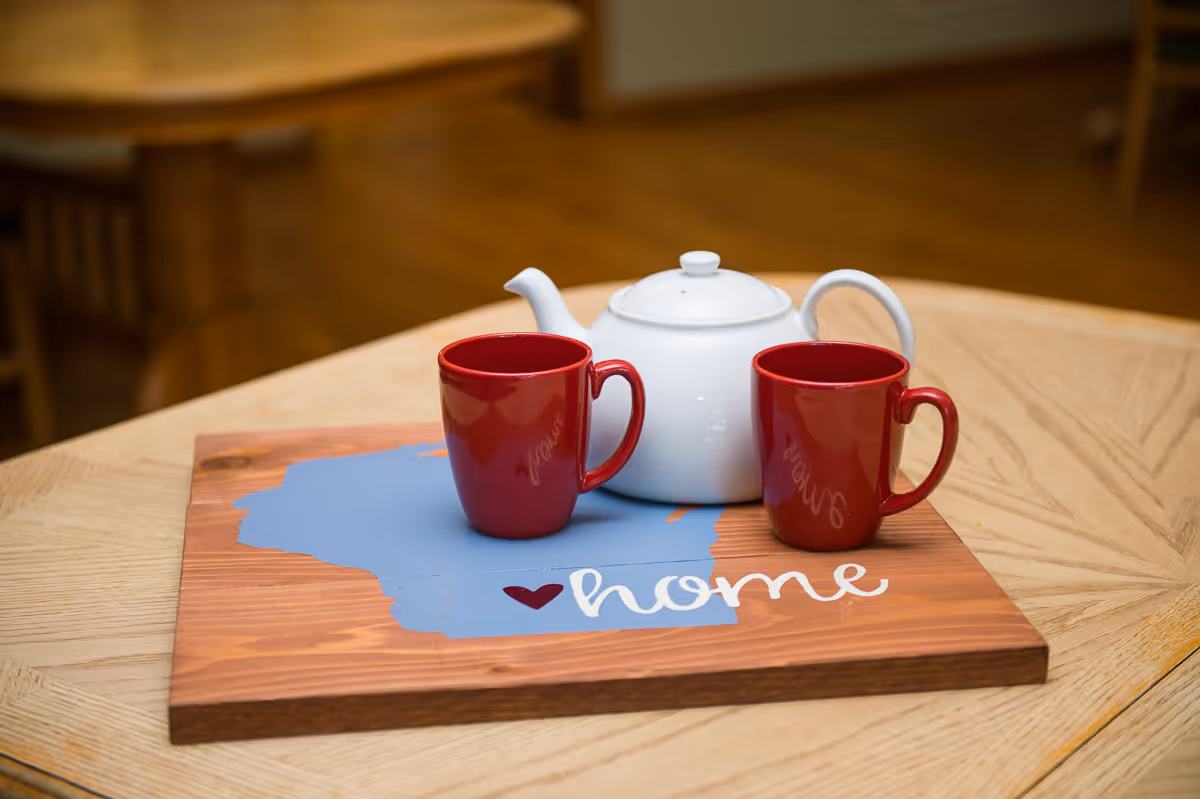 White teapot and two red mugs on a wooden tray with a blue shape and the word home on a light wood table.