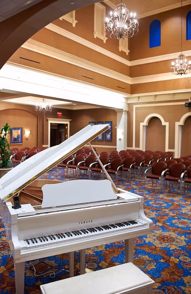 White Yamaha grand piano in an ornate event hall with rows of chairs, chandeliers, and patterned carpet.