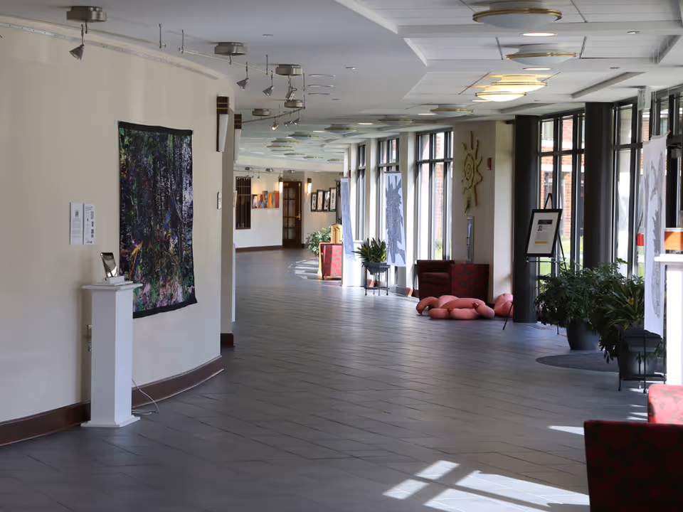 A long, bright hallway in a senior living facility with large windows on one side letting in natural light. The hallway features various pieces of artwork displayed on the walls and stands, along with some potted plants and comfortable seating areas with red chairs. The floor is tiled and the ceiling has multiple light fixtures.