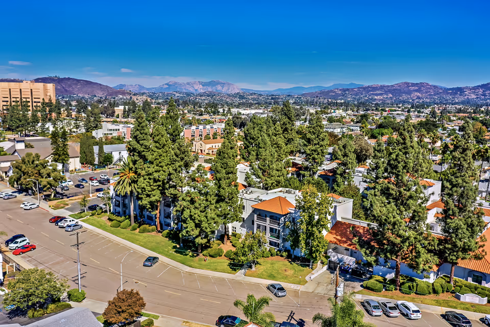 Aerial view of a senior living facility surrounded by tall trees and greenery, with a parking lot and several cars visible. The facility has white buildings with red-tiled roofs, set against a backdrop of distant mountains under a clear blue sky.