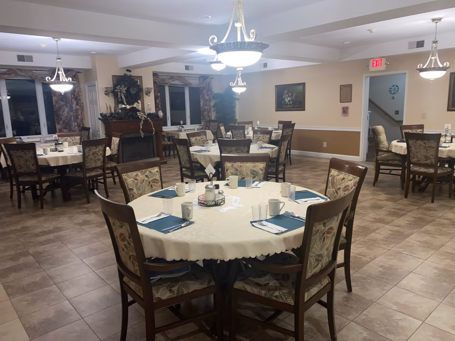 A dining room with several round tables covered with white tablecloths, each set with napkins, cups, and utensils. The chairs have floral patterned upholstery. The room has tiled floors, beige walls with a white chair rail, and multiple hanging light fixtures. There is a fireplace decorated with a wreath and other ornaments, and framed artwork on the walls. An exit sign is visible above a doorway.