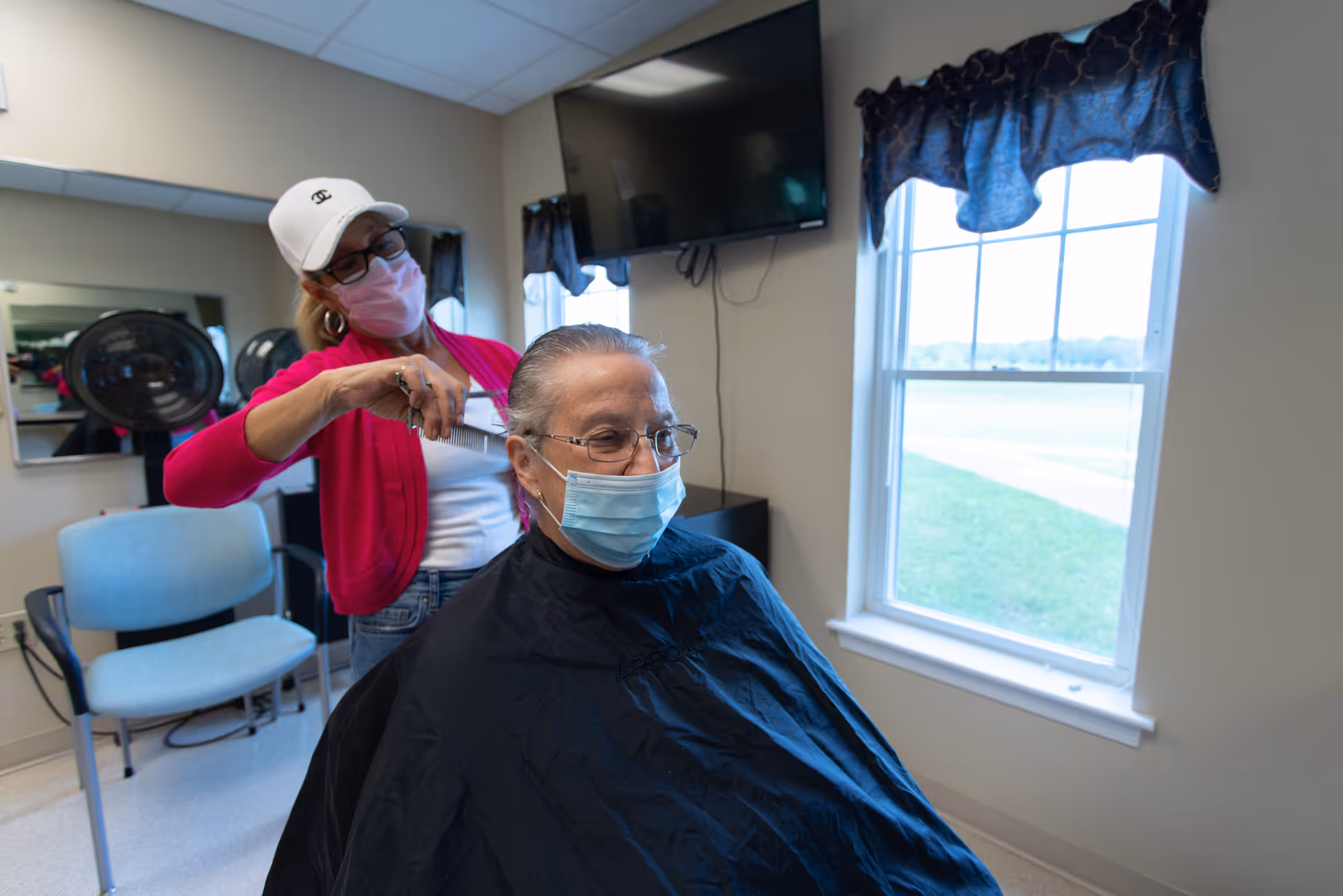 A woman wearing a white cap, glasses, and a pink face mask is cutting the hair of an elderly woman who is also wearing glasses and a blue face mask. They are in a well-lit room with a window showing an outdoor view, a TV mounted on the wall, and salon chairs in the background.