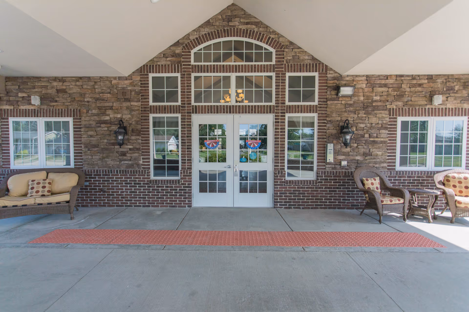 Entrance of a building with double glass doors framed by multiple windows and brick and stone exterior walls. There are outdoor wicker chairs with cushions on either side of the entrance under a covered area.