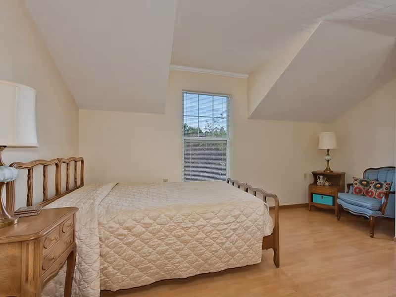 A bedroom with a wooden bed frame and a beige quilted bedspread. There is a wooden nightstand with a lamp on the left side of the bed and another wooden nightstand with a lamp and decorative items on the right side near a blue upholstered chair with colorful pillows. A window with blinds is centered on the far wall, letting in natural light. The room has light-colored walls and wooden flooring.
