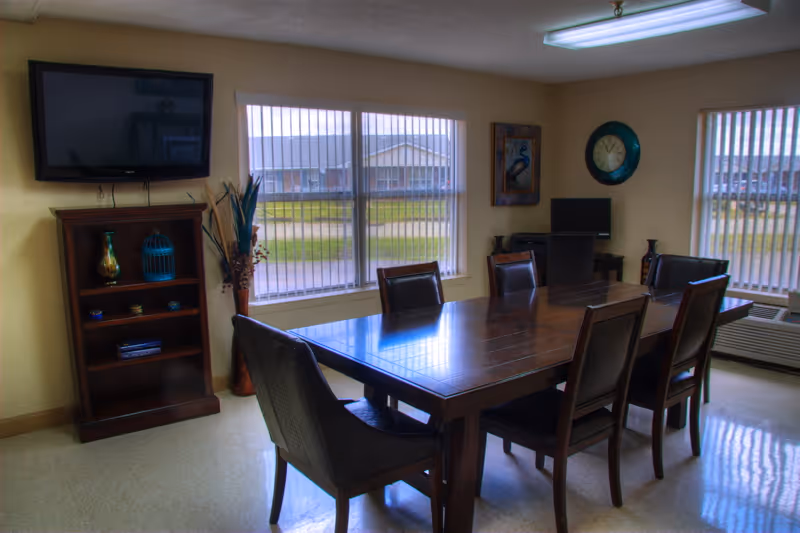 Communal dining room with a large wooden table surrounded by chairs, wall-mounted TV, bookshelf, and windows with vertical blinds.
