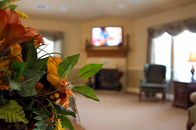 Close-up of a floral arrangement with green leaves and orange flowers in the foreground, with a blurred living room in the background featuring a fireplace, a mounted TV, an armchair, a window with curtains, and a side table with a lamp.