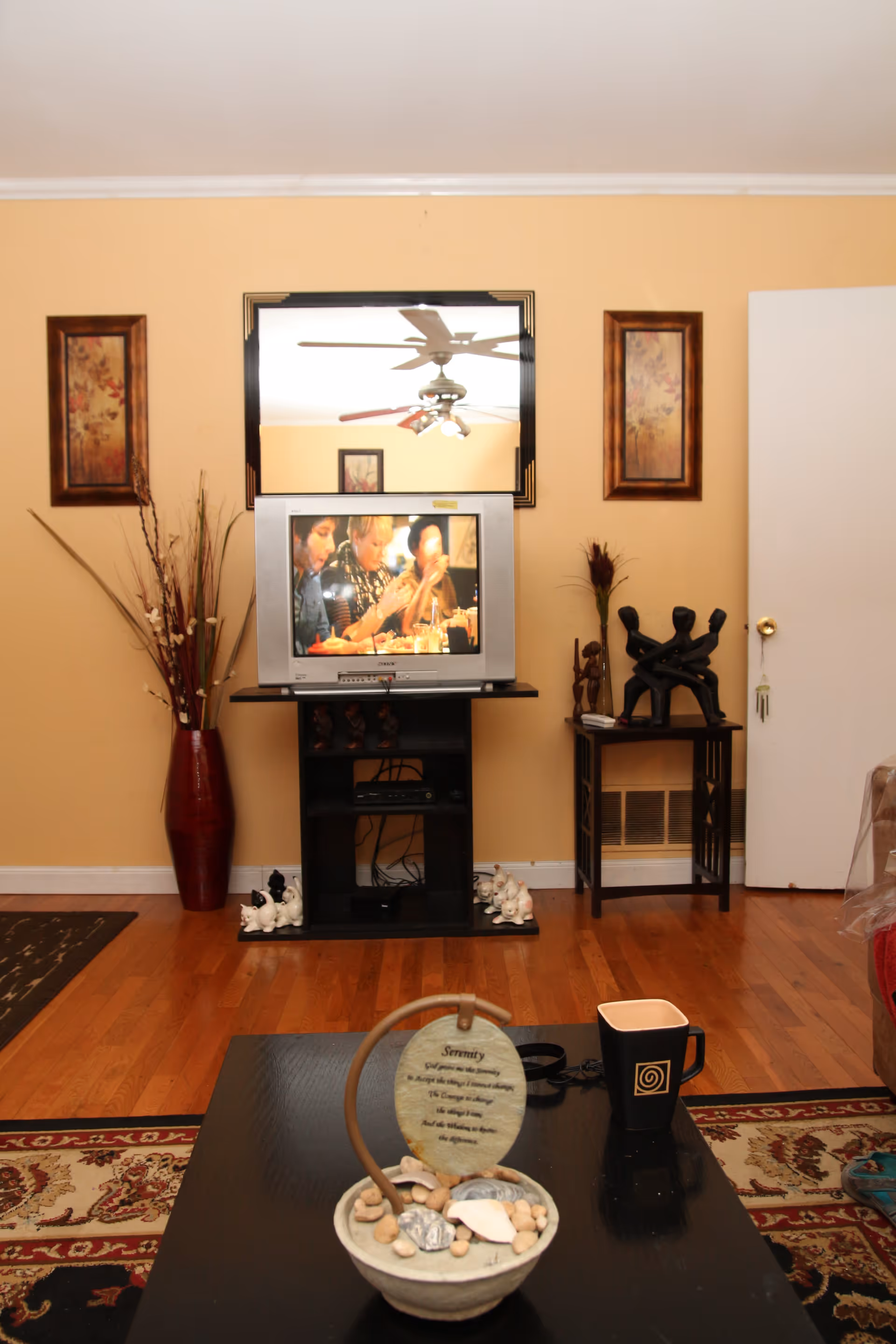 A cozy living room with a wooden floor, a TV on a black stand showing people dining, a large mirror above the TV reflecting a ceiling fan, two framed floral paintings on the wall, a tall red vase with decorative branches, a small side table with a black sculpture of three figures, and a coffee table with a decorative item and a black mug.