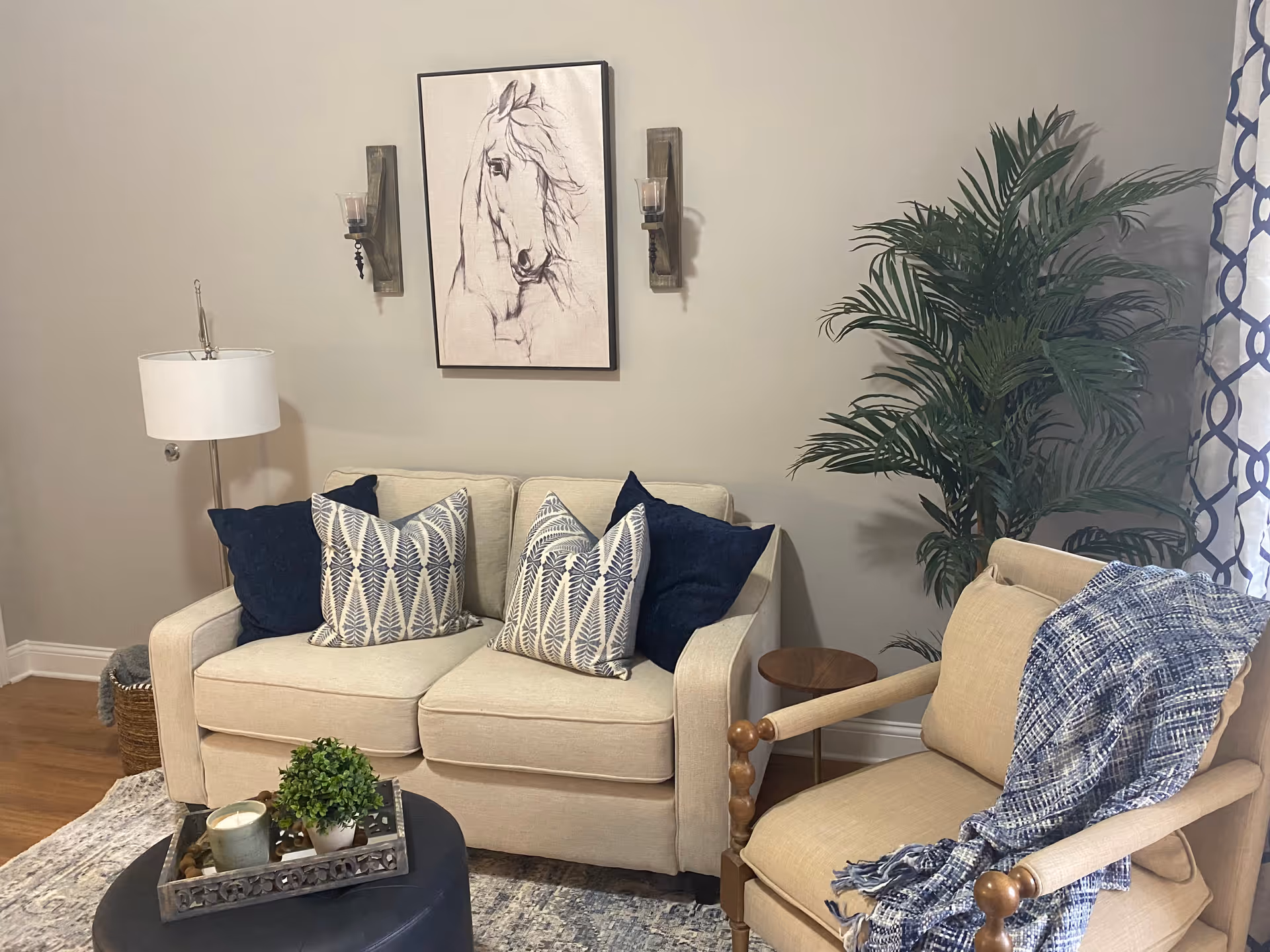 A cozy living room area with a beige loveseat adorned with four decorative pillows in navy blue and white patterns. To the right is a beige armchair with wooden armrests, draped with a blue and white throw blanket. A large green potted plant is placed in the corner near a window with patterned curtains. On the wall above the loveseat is a framed sketch of a horse's head flanked by two wall-mounted candle holders. A floor lamp with a white shade stands to the left of the loveseat, and a round black ottoman with a decorative tray holding a small plant and candle is in front of the seating area.