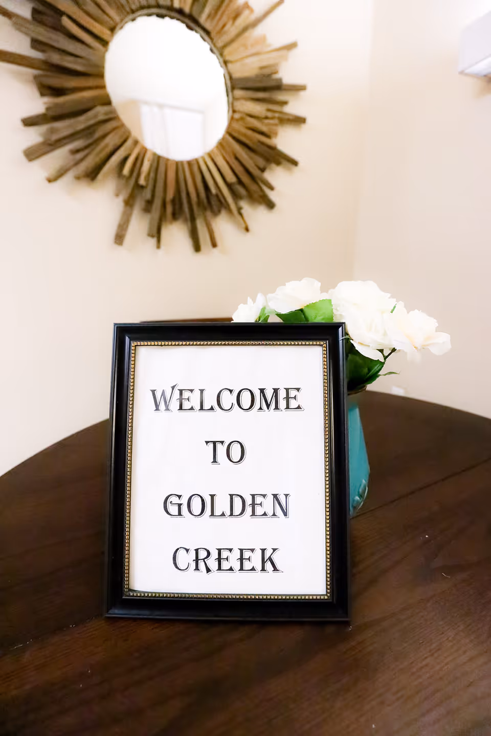A framed sign on a wooden table that reads 'WELCOME TO GOLDEN CREEK' with a vase of white flowers behind it and a decorative round mirror on the wall.