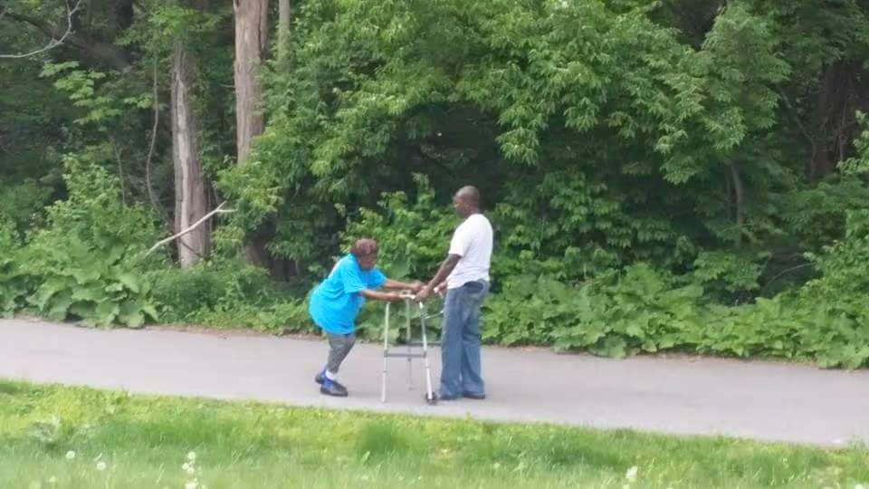 An elderly woman using a walker is assisted by a man on a paved path surrounded by lush green trees and grass.