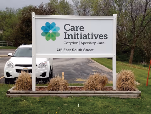 Outdoor sign reading "Care Initiatives Corydon | Specialty Care" with the address 745 East South Street in front of a parking lot and a parked white car.