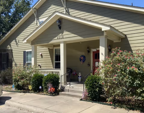Exterior view of a single-story residential building with beige siding and a small covered porch. The porch has a red door, two black lantern-style lights, and decorative items including a patriotic wreath and a small figure. The house number 4308 is displayed above the porch. There are bushes and flowering plants along the front of the house and a concrete driveway or sidewalk in front.