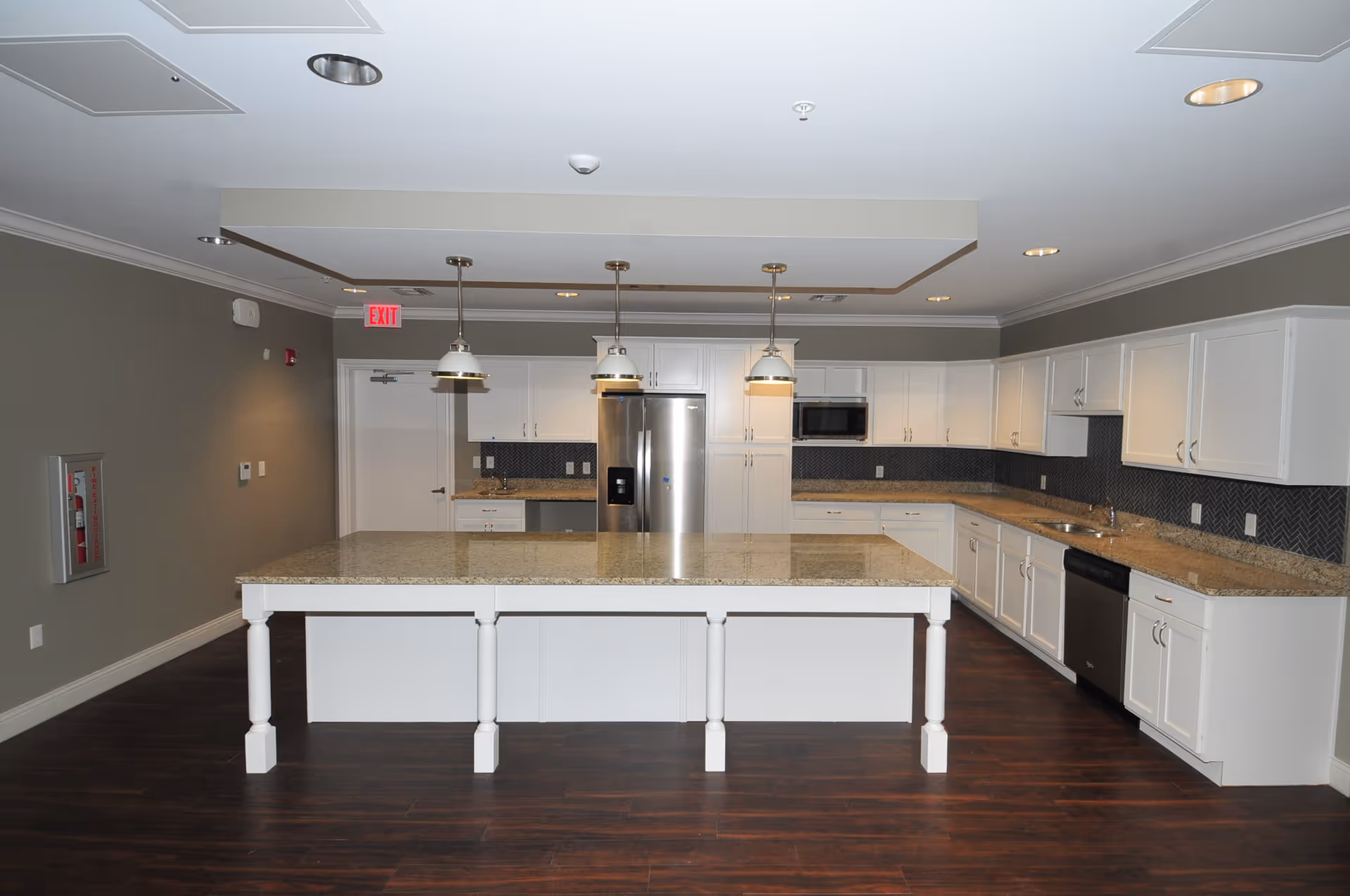 A modern kitchen with white cabinets, granite countertops, and dark wood flooring. There is a large central island with three pendant lights hanging above it. Stainless steel appliances including a refrigerator, microwave, and dishwasher are visible. The walls are painted gray and there is a fire extinguisher mounted on the left wall.
