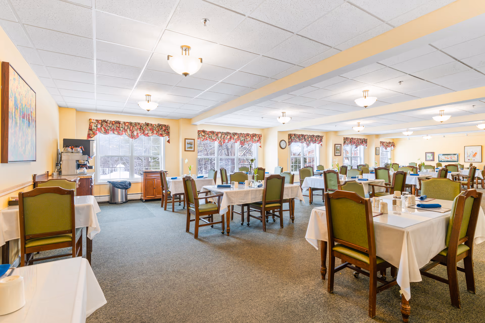 A bright and spacious dining room with multiple tables covered in white tablecloths, each set with cups, napkins, and utensils. The chairs have green upholstery and wooden frames. Large windows with floral valances let in natural light, and the walls are painted a soft yellow. Ceiling lights provide additional illumination. There is a coffee station and trash bin near the windows.