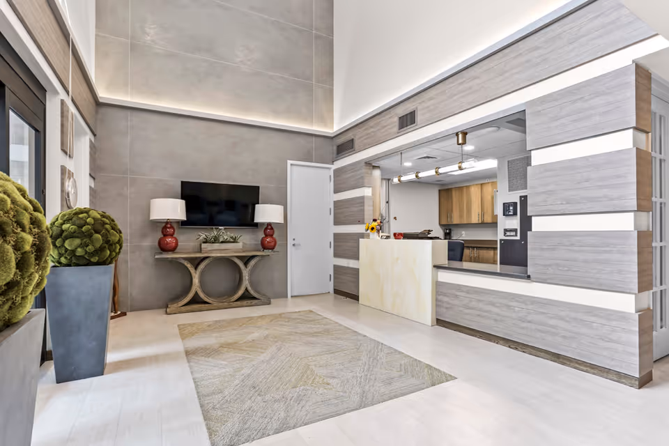 Modern interior lobby with a reception desk, a wall-mounted TV above a console table flanked by red lamps, decorative plants, and a rug.
