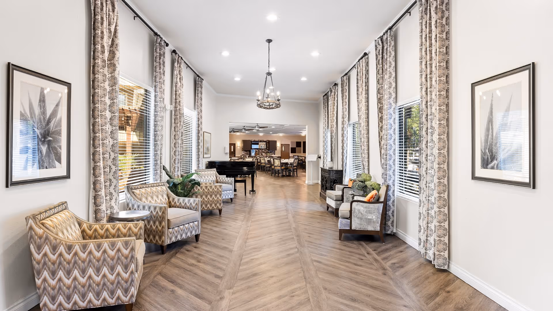 A bright and spacious interior hallway with large windows on both sides covered by patterned curtains. The hallway features several upholstered armchairs arranged along the walls, a chandelier hanging from the ceiling, and framed botanical artwork on the walls. At the far end, there is a view into a dining area with tables and chairs, and a black grand piano is positioned near the windows on the left side.