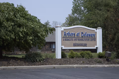 A large entrance sign reading "Arbors at Delaware" set in a landscaped bed with shrubs and the facility building visible behind it.