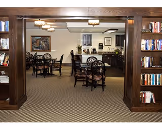 Interior view of a dining area in a senior living facility with multiple round tables and chairs. The room features dark wood bookshelves on either side of the entrance, a patterned carpet, and a kitchenette area in the background with cabinets, a countertop, and a coffee station.