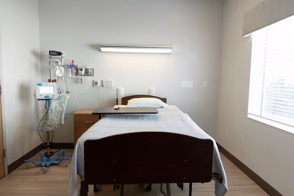 A hospital-style room with a single bed covered in white linens and a pillow. There is a bedside table with medical equipment attached to the wall and a medical monitor on a stand next to the bed. A large window with blinds is on the right side, allowing natural light into the room. The walls are plain and light-colored, and the floor has a wood-like finish.