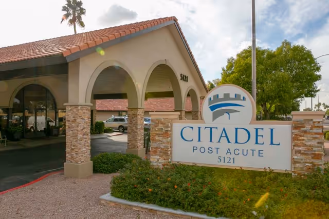 Exterior view of the Citadel Post Acute facility showing a building with arched entrance supported by stone pillars, a tiled roof, and a large sign in front with the facility name and address 5121. There are some bushes and trees around the sign and parking area visible in the background.