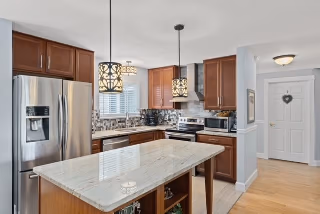 A modern kitchen with wooden cabinets, a large marble island in the center, stainless steel refrigerator, stove, dishwasher, and microwave. Two decorative pendant lights hang above the island, and there is a window with blinds above the sink. The floor is light wood, and a white door is visible in the background.