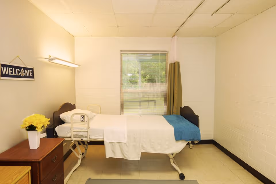 Simple single-occupancy care facility bedroom with a hospital-style bed, window, nightstand with flowers and a 'Welcome' sign.