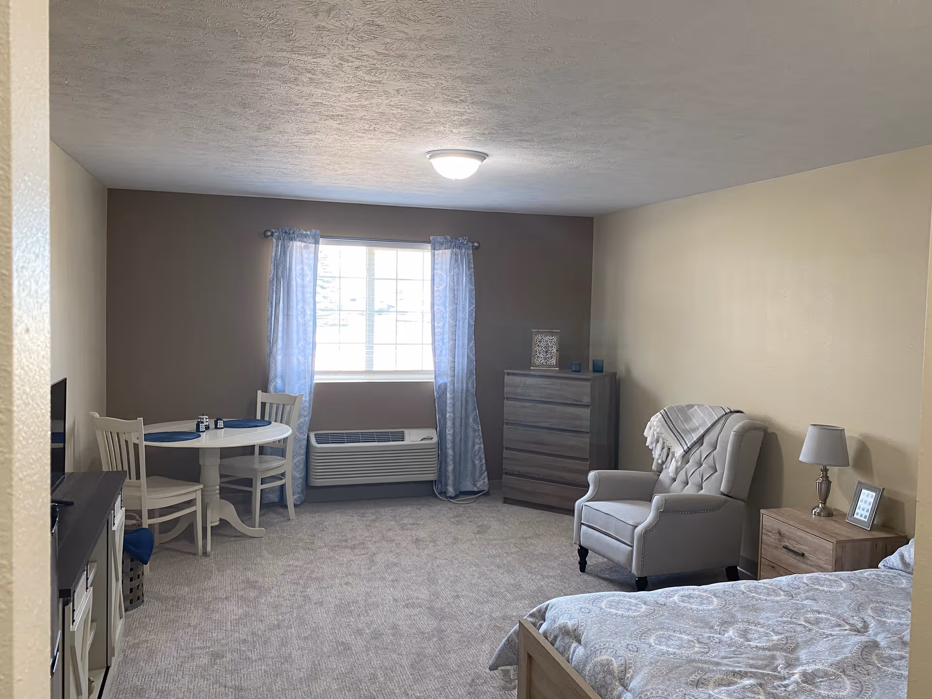A cozy living space with a bed partially visible in the foreground, a cushioned armchair with a throw blanket, a wooden nightstand with a lamp and picture frame, a tall dresser, a small round dining table with two chairs, and a window with light blue curtains letting in natural light. The room has beige walls and carpeted flooring.