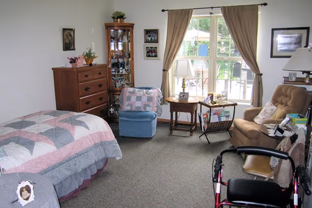 A cozy bedroom in an assisted living facility featuring a bed with a quilted blanket, a wooden dresser, a glass display cabinet, a blue armchair with a quilt draped over it, a side table with a lamp, a window with beige curtains, a brown recliner chair, and a walker in the foreground.